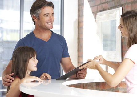 A female receptionist handing a father some paperwork while he smiles back and stands next to his daughter